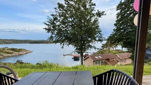 A wooden deck with a table and chairs, overlooking a calm lake with a dock and houses in the background. - 5 Star Holiday Home in Uddevalla (Uddevalla)