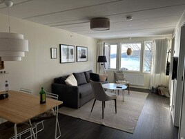 Una sala de estar moderna con un sofá, una mesa de comedor y una mesa de centro. Hay cuadros enmarcados en la pared, una lámpara y vistas al exterior a través de grandes ventanales.