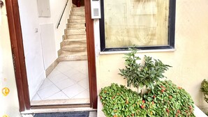 L’entrée d’un immeuble avec une porte en bois, un escalier, une boîte aux lettres et un petit jardin agrémenté de fleurs rouges.
