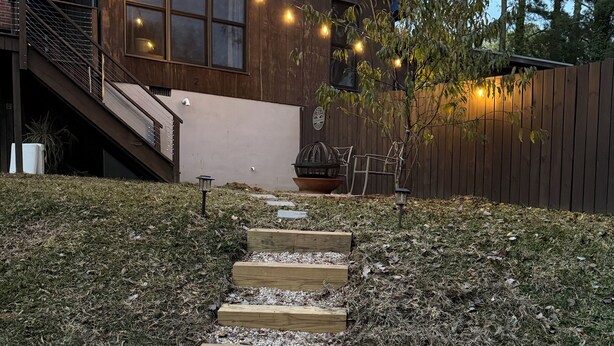 Ein Holzhaus mit einer Treppe zu einer Terrasse, umgeben von einem Zaun und Bäumen.