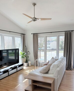 A modern living room with a flat-screen TV, a sofa, a ceiling fan, and large windows. - Cozy Studio Downtown Raleigh (Raleigh)