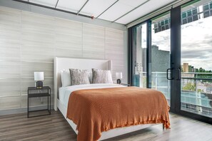 Et moderne hotelværelse med en stor seng, et natbord, en lampe og udsigt over byen gennem en glasdør.