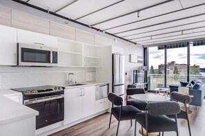Et moderne køkken med indbygget ovn, mikroovn og køleskab, et spiseområde med et rundt bord og stole, samt store vinduer med udsigt over byen.