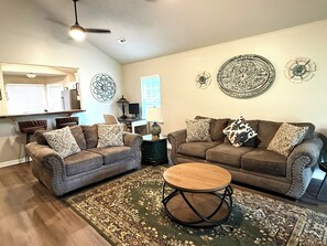 A living room with two sofas, a coffee table, decorative wall art, a ceiling fan, and a window with blinds. - Spacious Home Near HSV Airport & Entertainment - Space & Rckt, Orion amp theatre (Madison)