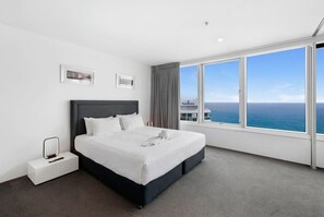 Une chambre d’hôtel moderne avec un grand lit, une table de chevet et une vue sur la mer, avec un paysage urbain en arrière-plan.