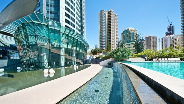 A modern building with a glass facade, a pool area with clear water, and high-rise buildings in the background. - Oversized South-Facing Apartment | Q1 Level 39 (Surfers Paradise)