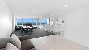 A modern living room with a sofa, a dining area with a table and chairs, a kitchen area, and large windows offering a view of the sea. - Oversized South-Facing Apartment | Q1 Level 39 (Surfers Paradise)