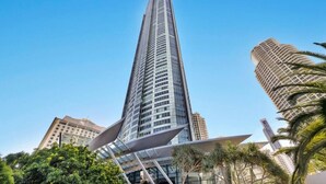 A tall, modern skyscraper with a unique angular design, surrounded by other buildings and palm trees. - Oversized South-Facing Apartment | Q1 Level 39 (Surfers Paradise)