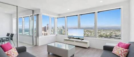 A modern living room with a glass coffee table, grey sofas, a flat-screen TV, and large windows offering a city view.