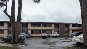 Un aparcament amb diversos cotxes, un edifici amb façana beig i arbres al primer pla.