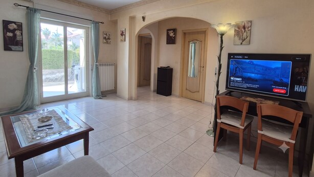 Uma sala de estar com televisão, duas cadeiras de madeira, uma mesa de centro de vidro e uma porta de correr que dá para o exterior.