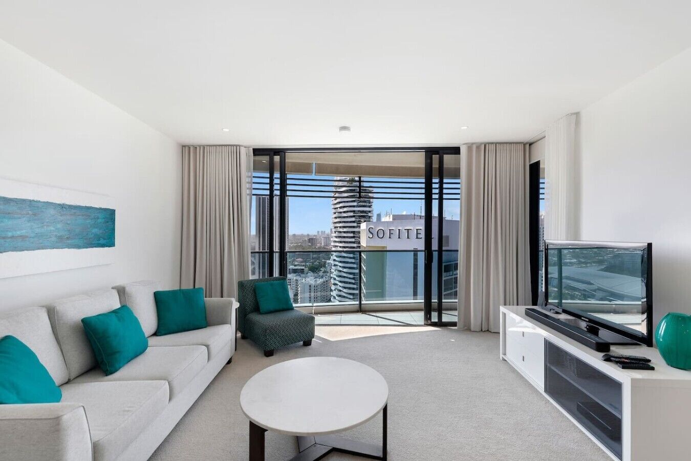 Ett modernt hotellrum med en soffa, ett litet bord, en TV och en balkong med utsikt över staden.