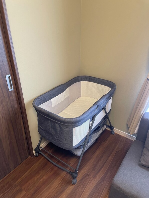 A corner baby crib with a grey frame and white mattress, positioned against a wall with a wooden floor.