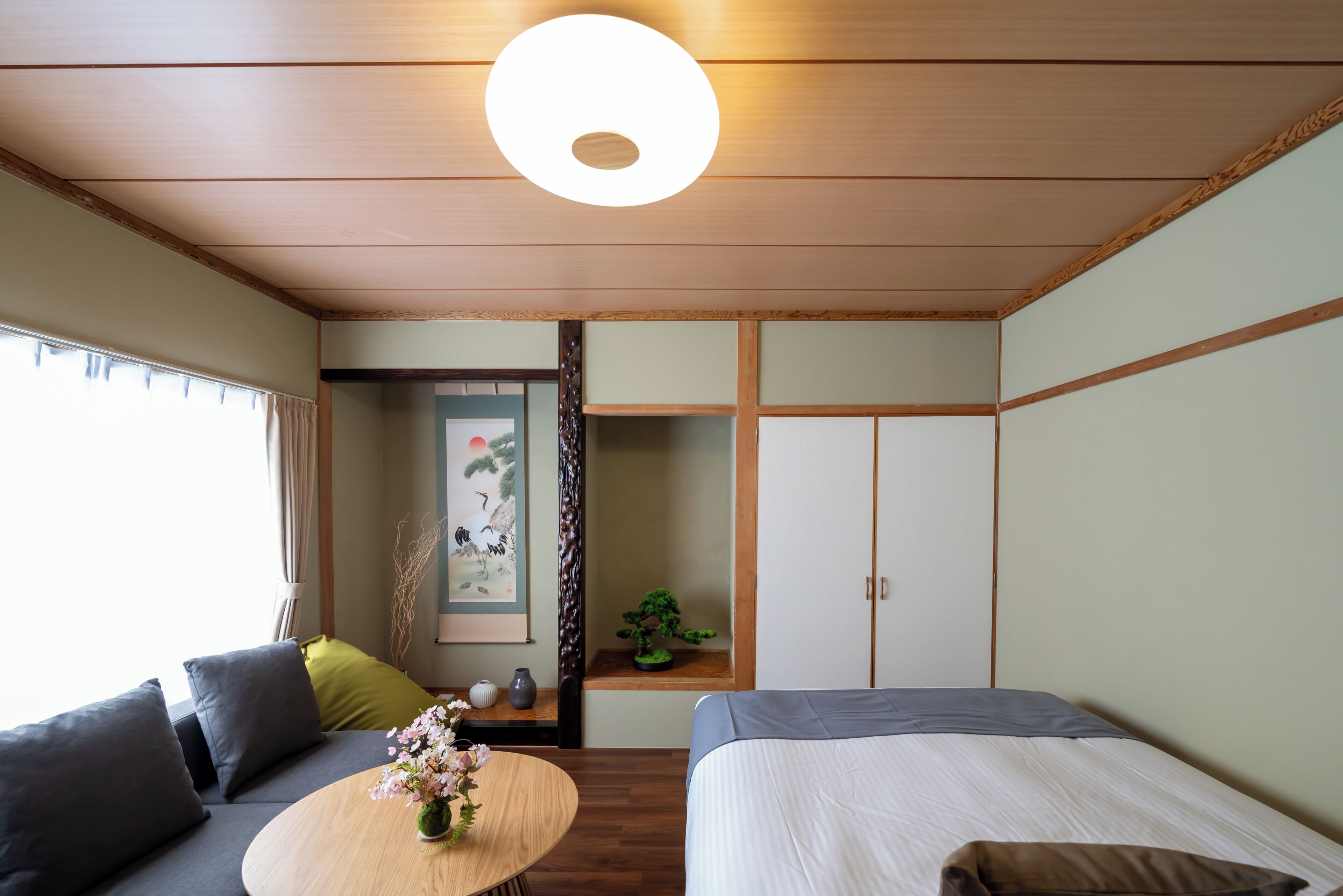 A room with a bed, a sofa, a small table with a vase, a wall-mounted shelf with a plant, and a traditional Japanese sliding door.