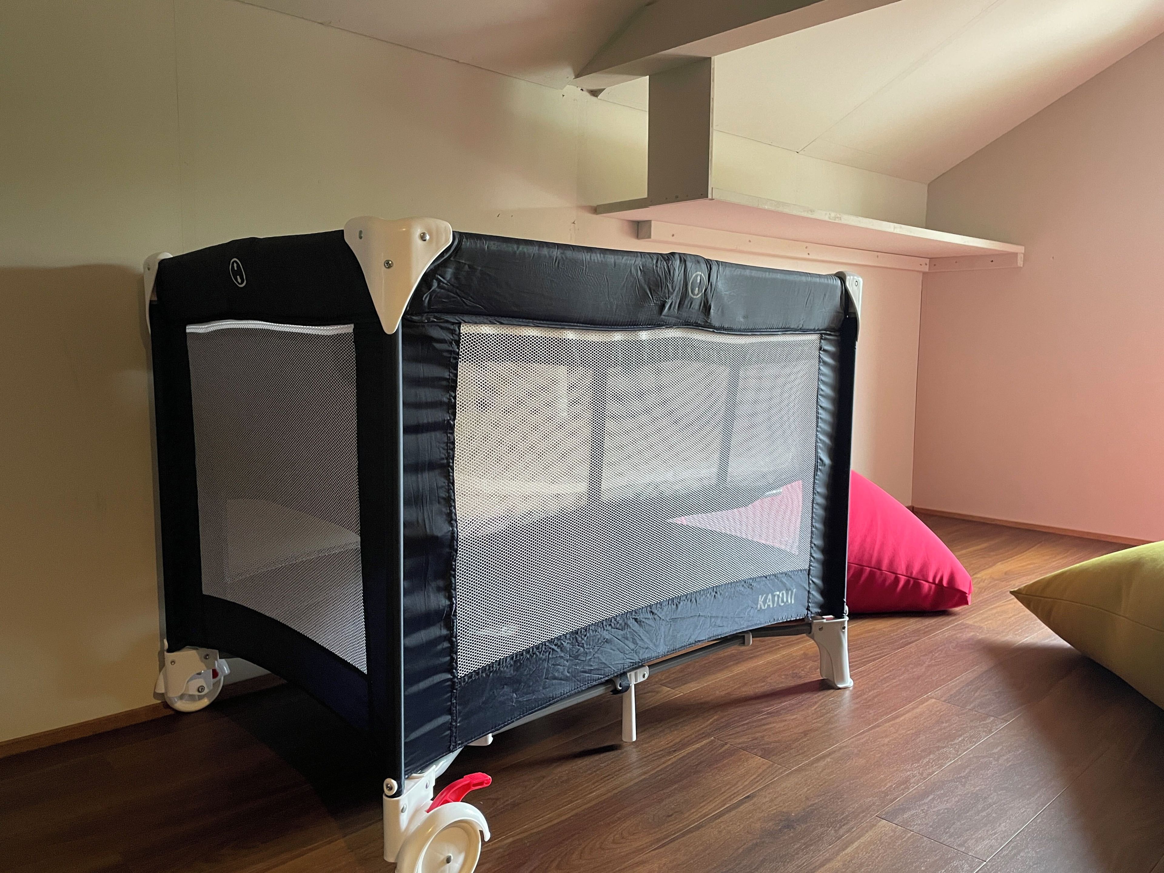 A foldable playpen with wheels, a red cushion, and a wooden floor.