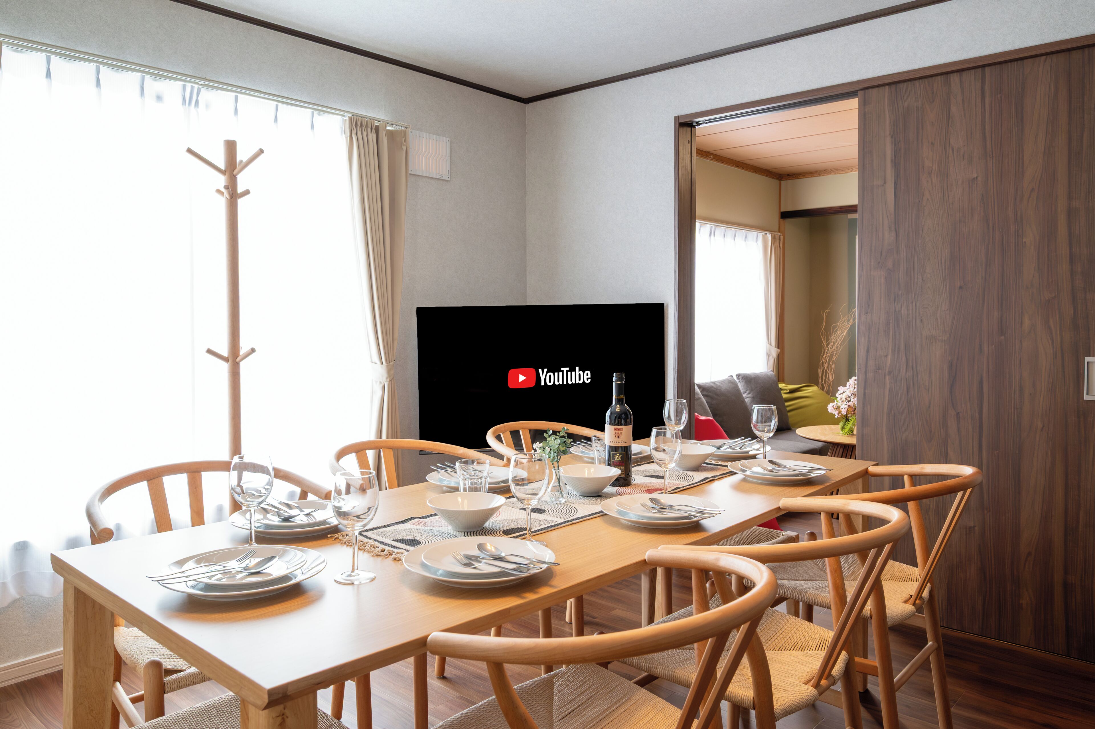 A dining area with a wooden table set for four, a coat rack, and a television in the background.