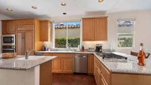 A modern kitchen with wooden cabinets, granite countertops, a built-in dishwasher, and a large window providing a view of the outdoors. - Little Sol at The Palms Golf Club (La Quinta)