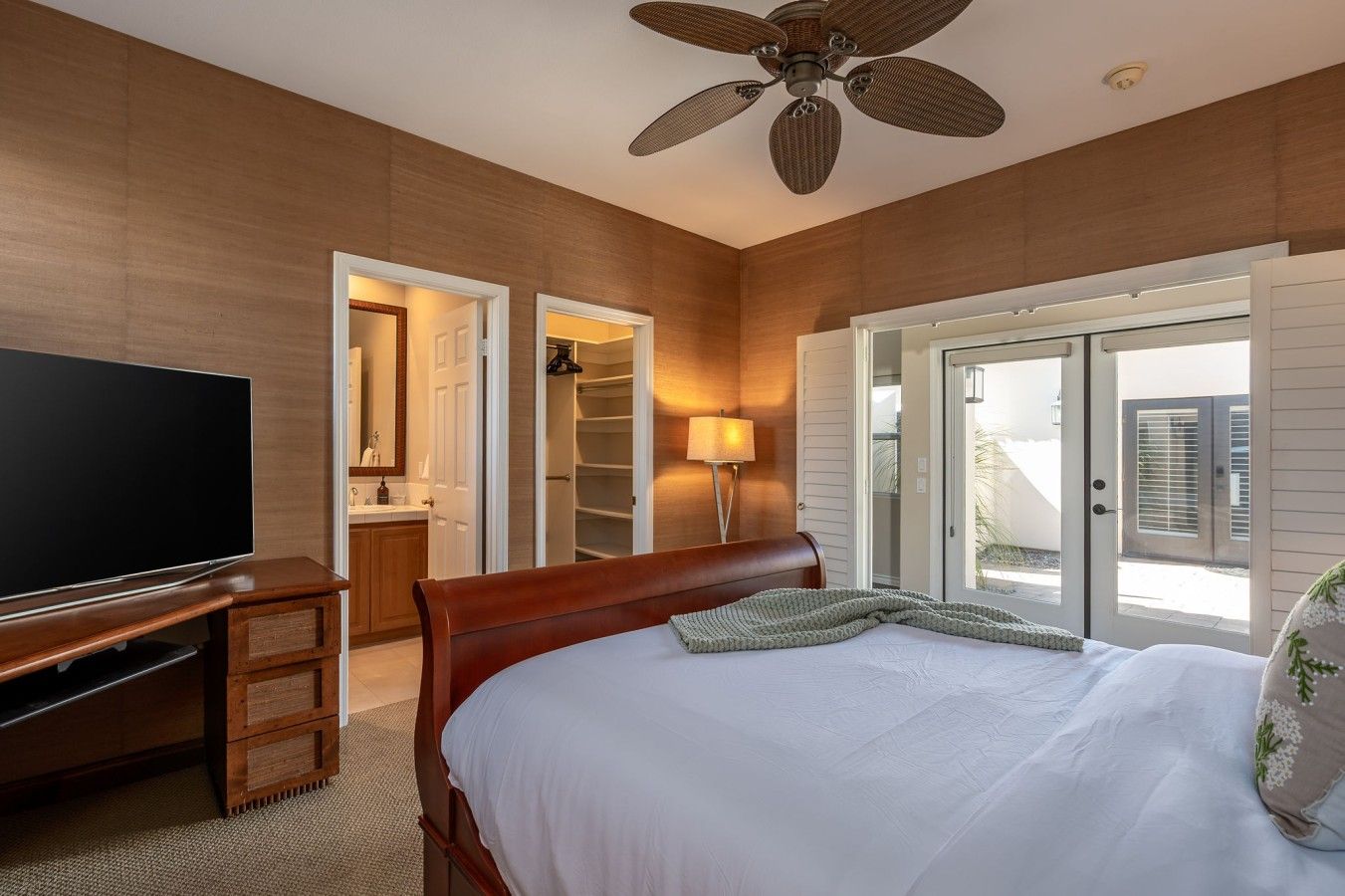 Quarto de hotel com cama, televisão, ventoinha no teto, closet amplo e vista através de uma porta de vidro.