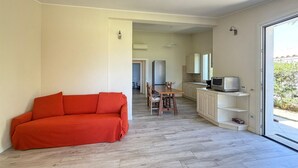 En moderne stue med en rød sofa, et spiseområde med bord og stole, et køkken med skabe og en mikrobølgeovn, samt en skydedør, der fører til et udendørs område.