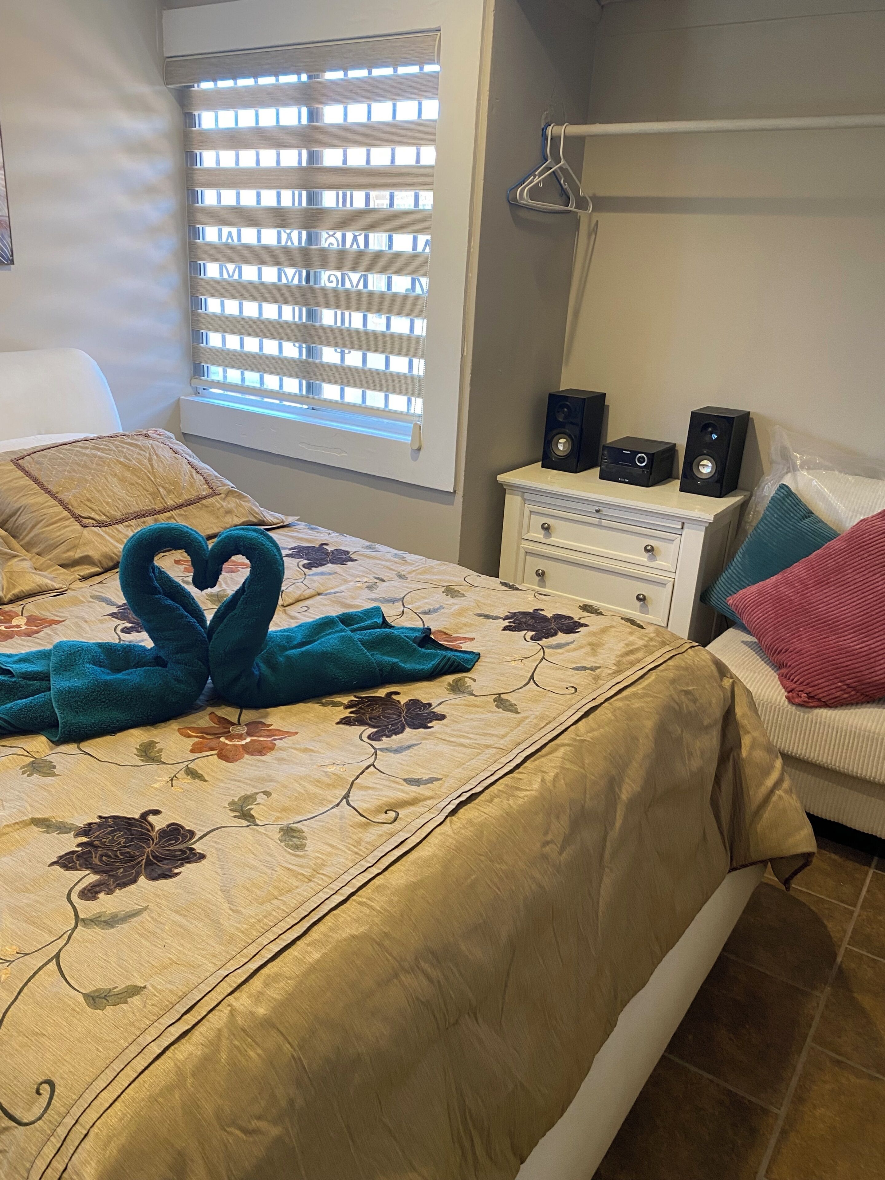 A bedroom with a bed, a dresser, a window with blinds, and two speakers on the dresser.