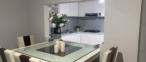 A modern dining area with a glass-top table, white chairs, and a kitchen in the background.