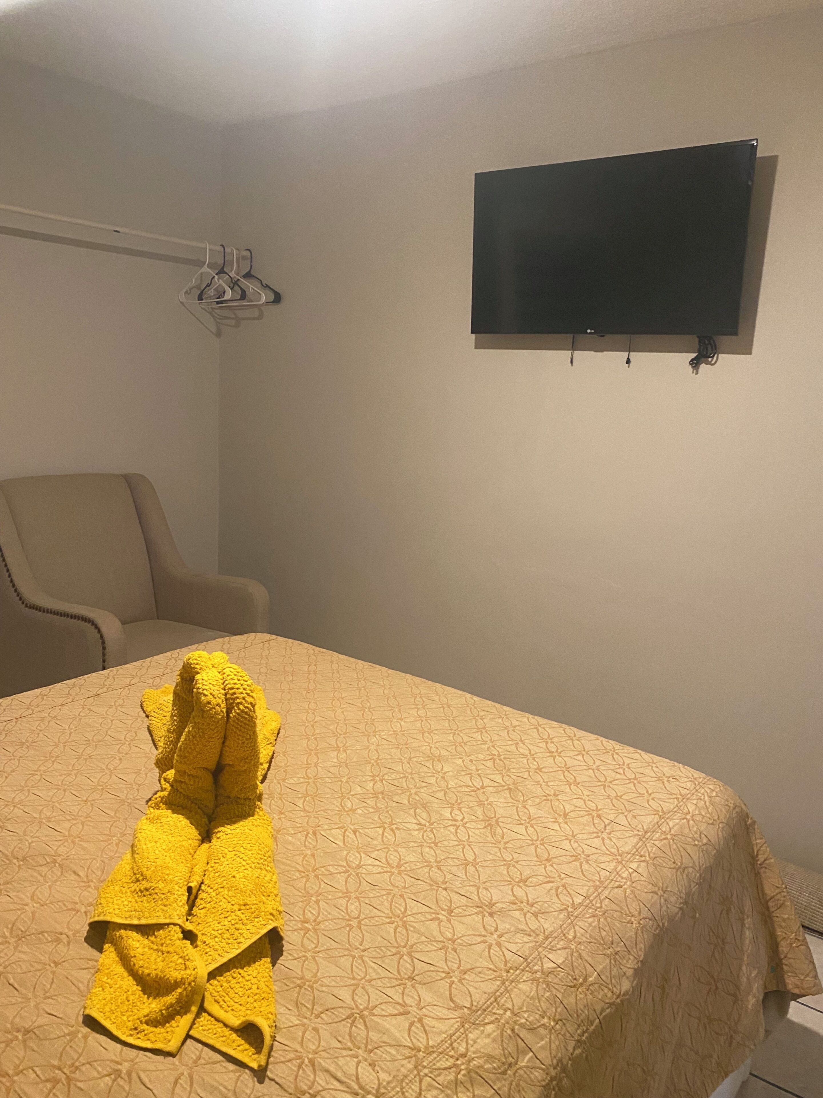 Hotellihuone, jossa on sänky, tuoli, seinälle asennettu televisio ja pyyhe sängyllä.