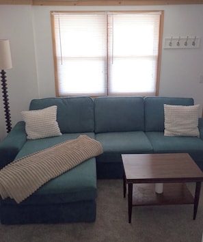Una sala de estar con un sofá verde, dos almohadas blancas, una manta color beige, una mesa de centro de madera y una ventana con persianas.
