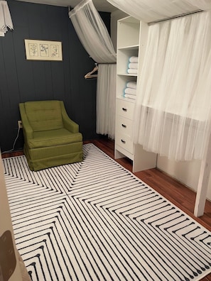 Una habitación con un sillón verde, un mueble blanco, un tapete estampado y una cama con dosel.