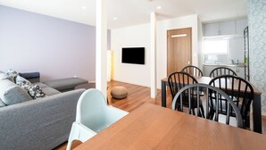 A modern living room with a sofa, a dining table with chairs, a flat-screen TV mounted on the wall, and a kitchen area with cabinets and a window. - Villa Shigin (Sorachi-gun,Nakafurano-cho)