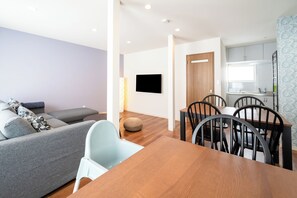 A modern living room with a sofa, a dining table with chairs, a flat-screen TV mounted on the wall, and a kitchen area with cabinets and a window. - Villa Shigin (Sorachi-gun,Nakafurano-cho)