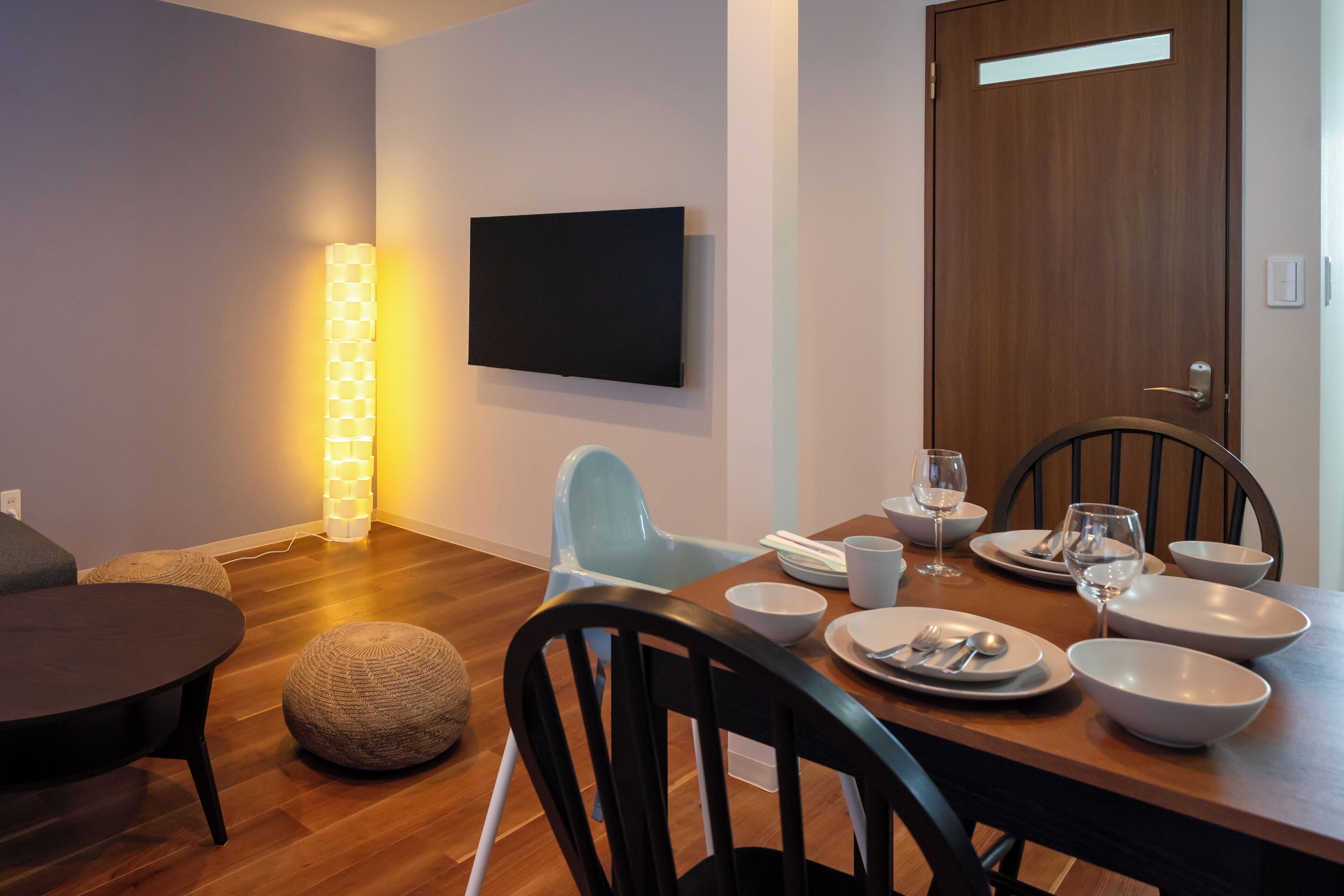 A modern dining area with a wooden table set for four, a flat-screen TV mounted on the wall, and a standing floor lamp.