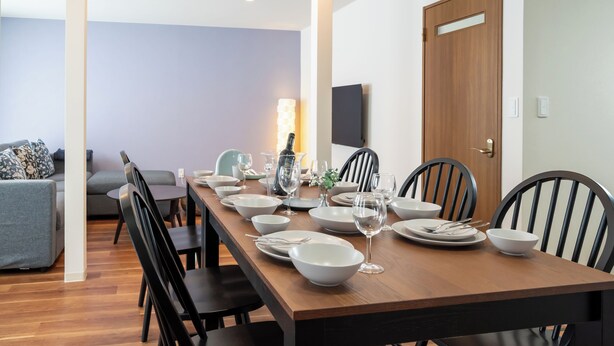 Un comedor con una mesa de madera alargada dispuesta para cuatro personas, rodeada de sillas negras, con un sofá gris y un televisor montado en la pared al fondo.