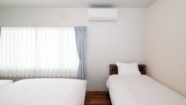 A hotel room with two beds, a window with sheer curtains, an air conditioning unit, and a wooden nightstand. - Villa Shigin (Sorachi-gun,Nakafurano-cho)