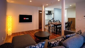 Una sala de estar moderna con un televisor de pantalla plana, un comedor con sillas y una mesa, y una cocina al fondo.