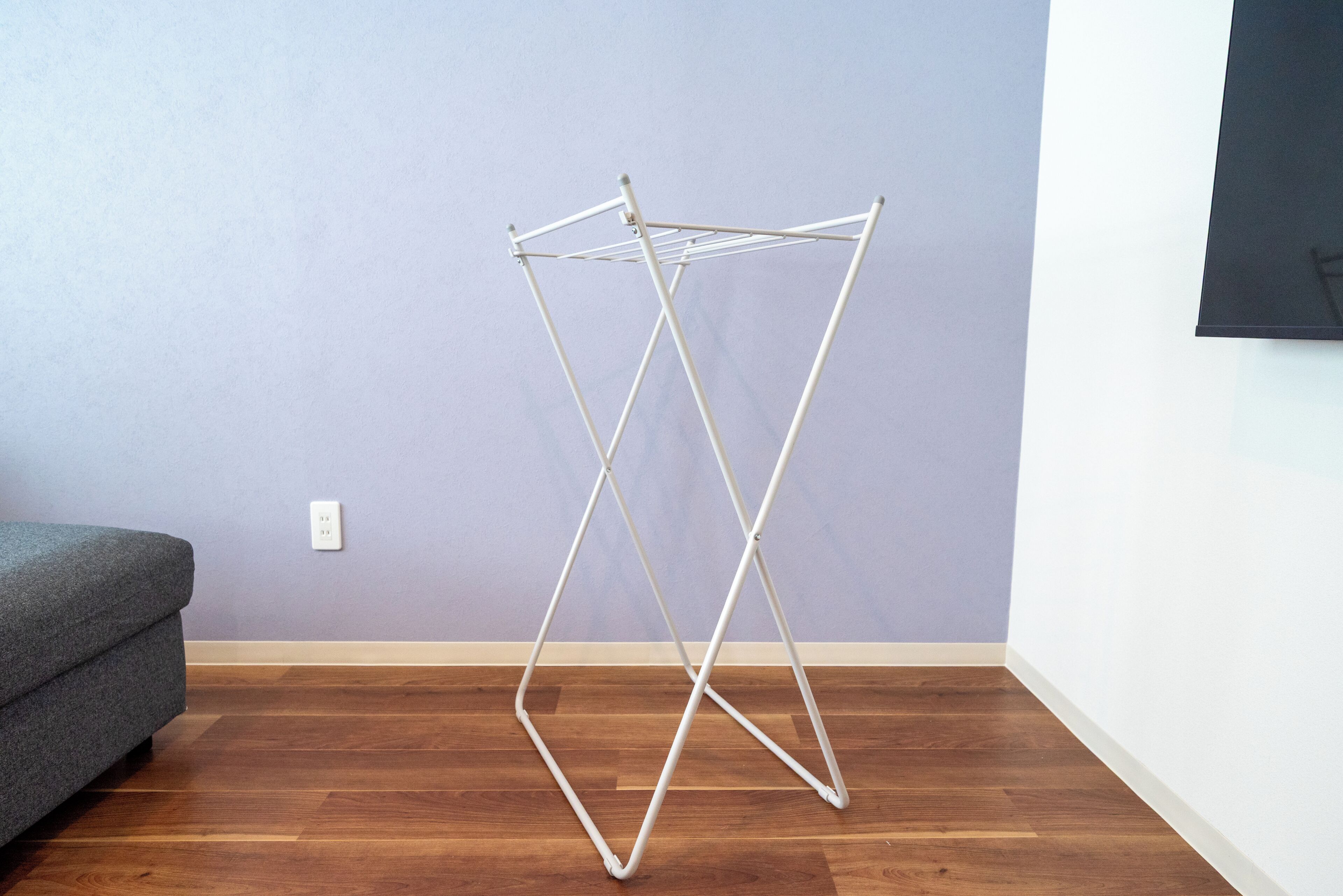 Une pièce avec un plancher en bois, un canapé gris et un étendoir à linge en métal blanc adossé à un mur violet.