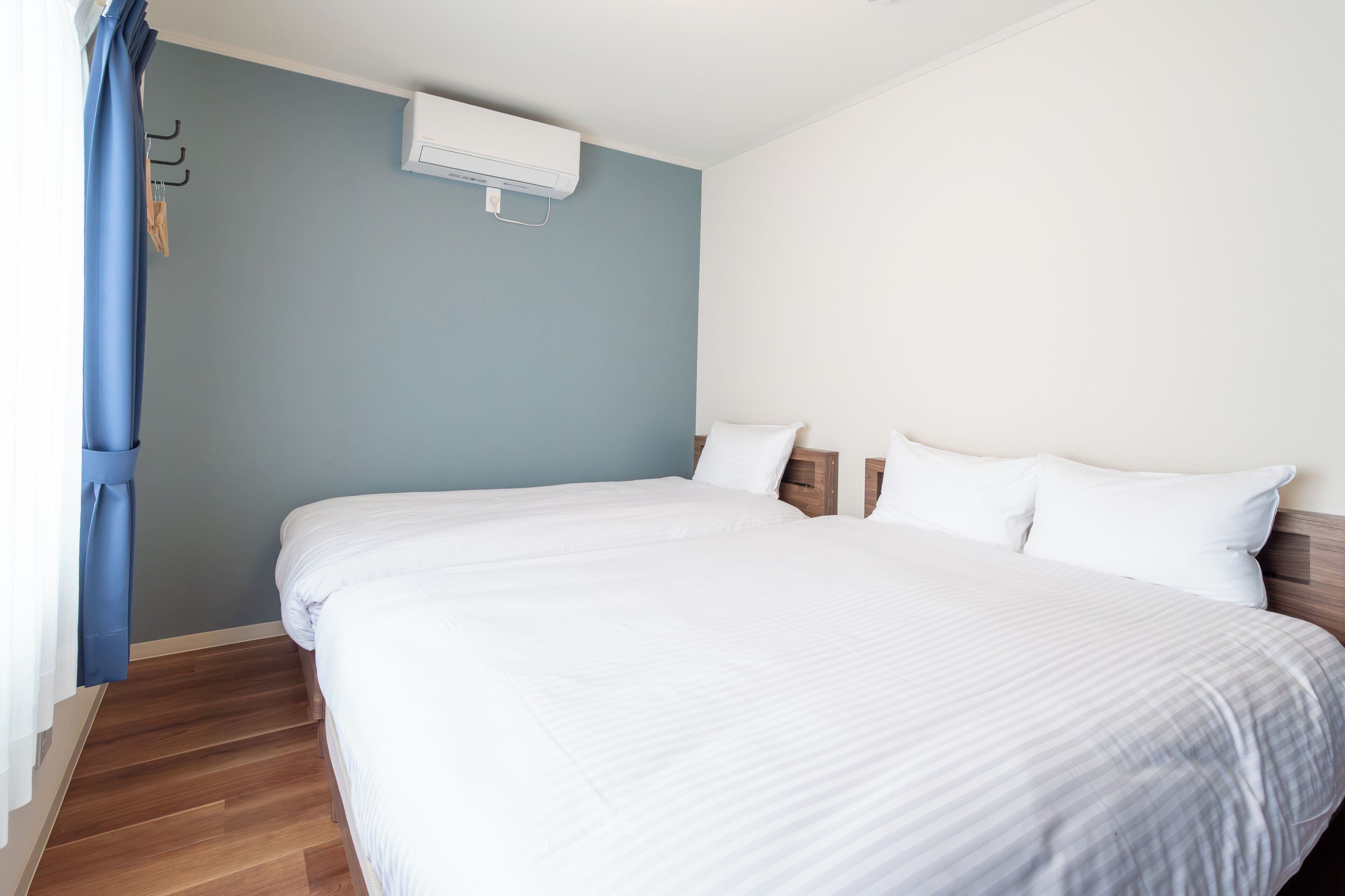 A neatly made bed in a room with a blue wall, a wooden floor, and a white air conditioning unit.