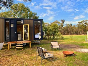 A modern, compact mobile home with a wooden deck, a dining table, two chairs, and a small outdoor seating area.
