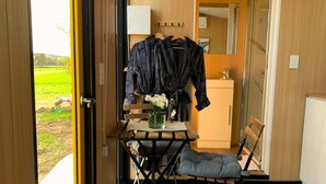 Una habitación con piso de madera, una mesa con una silla, un banco, una mesita con un jarrón de flores y un perchero con un abrigo.