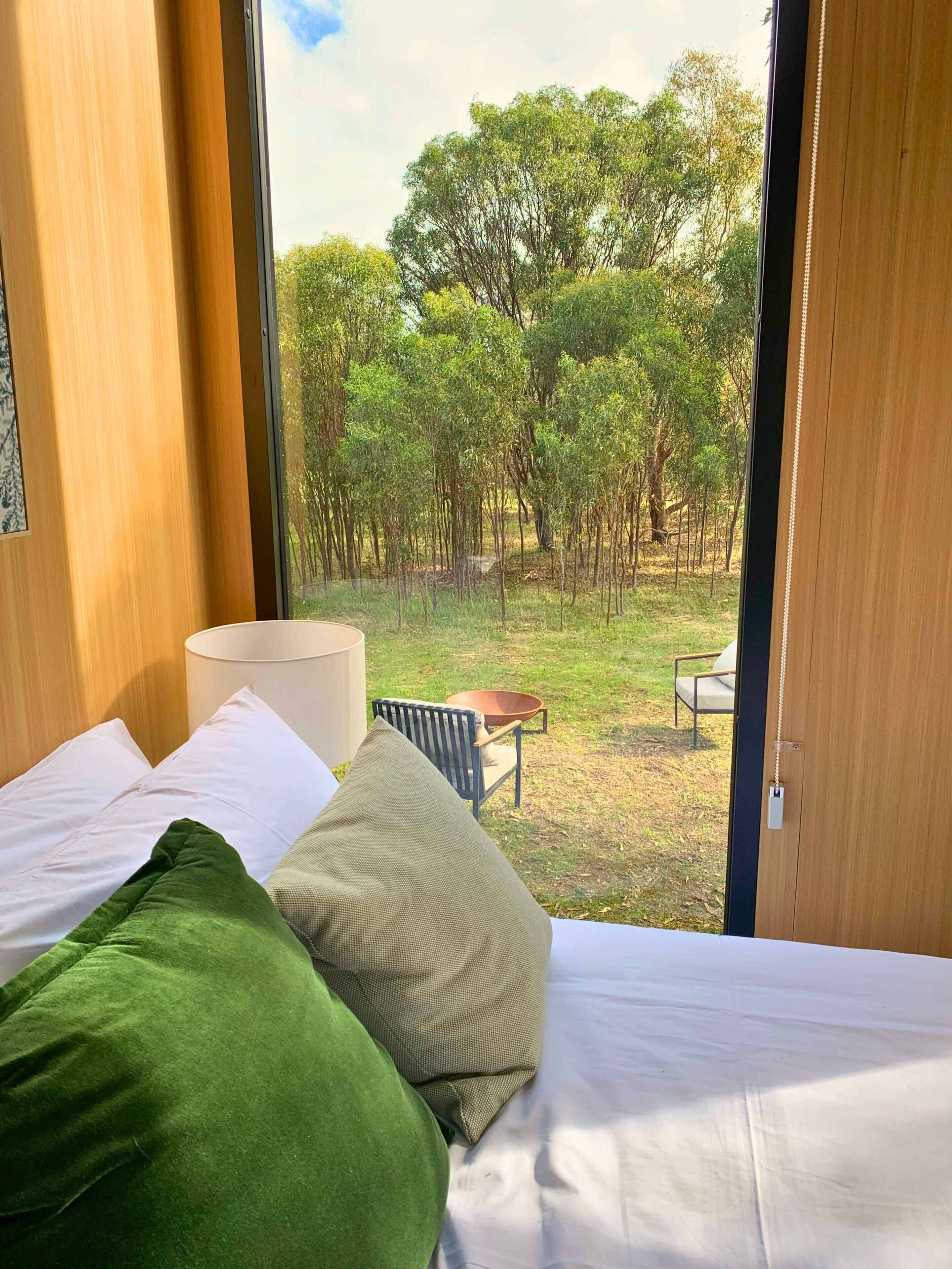 Un dormitorio con cama, almohadas, una silla y vista a un espacio verde con árboles.