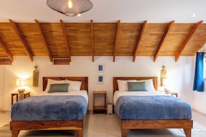 Quarto com duas camas de madeira, vigas de madeira no teto, uma pequena mesa e luminária fixada na parede.