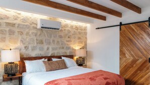 Ein Schlafzimmer mit Steinwand, Holzbalkendecke, einem Bett mit weißer Bettwäsche, zwei Nachttischlampen und einer Holztür mit Fischgrätmuster.