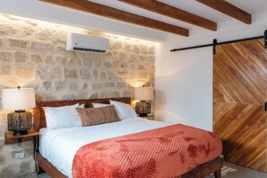 Um quarto com parede de pedra, vigas de madeira no teto, uma cama com roupa de cama branca, duas luminárias de cabeceira e uma porta de madeira com padrão em espinha de peixe.