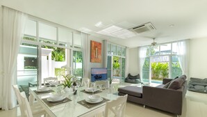 A modern living room with a glass dining table, white chairs, a grey sofa, and a TV. - Private Pool Villa Oxygen Bangtao 3BR (Tambon Choeng Thale)