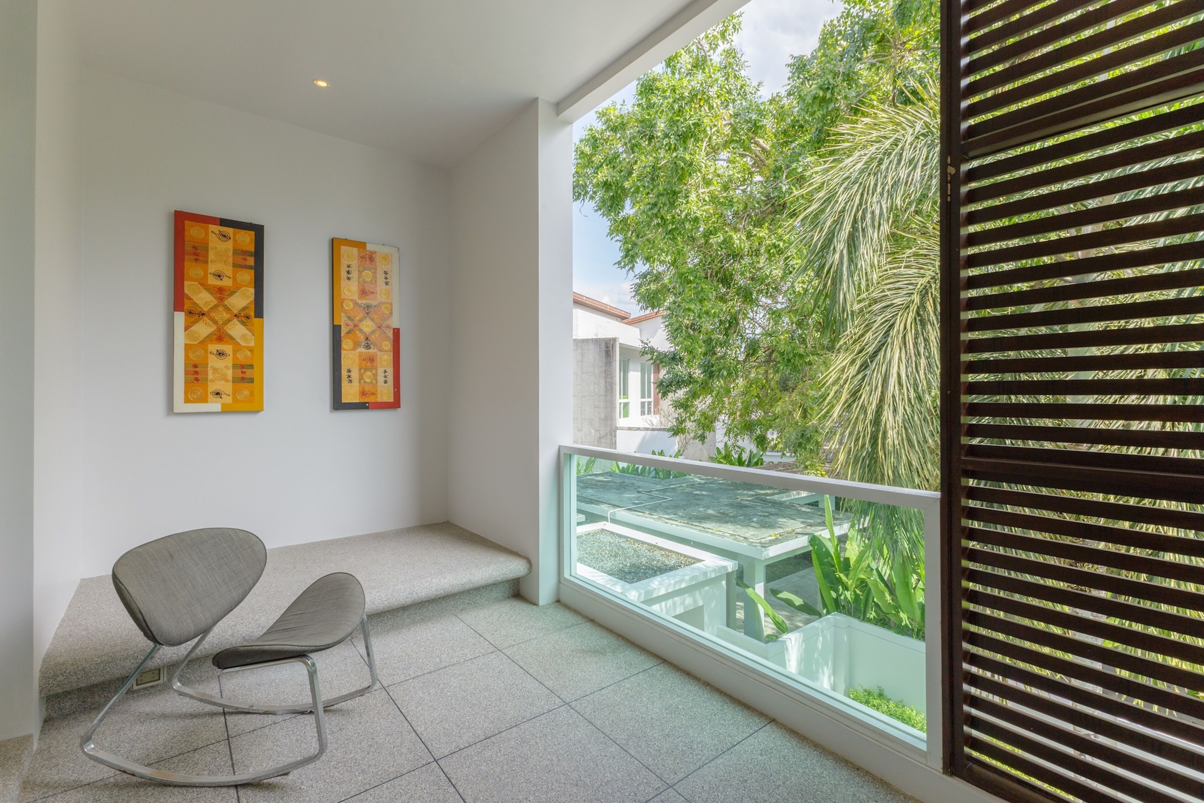 A balcony with a view of greenery, a seating area with chairs, and two artworks on the wall.