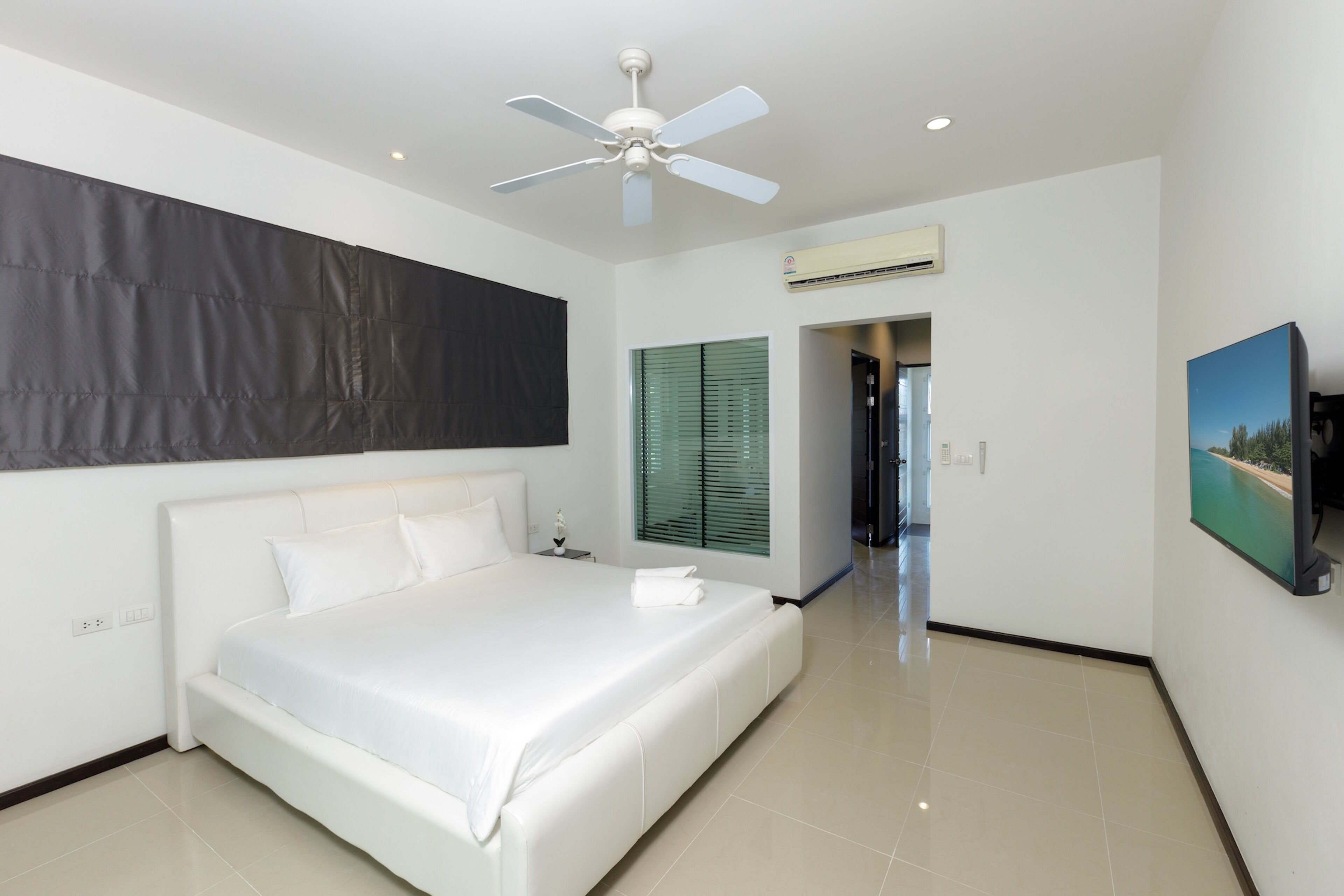 A modern bedroom with a large bed, a ceiling fan, an air conditioning unit, a painting, and a view into another room.