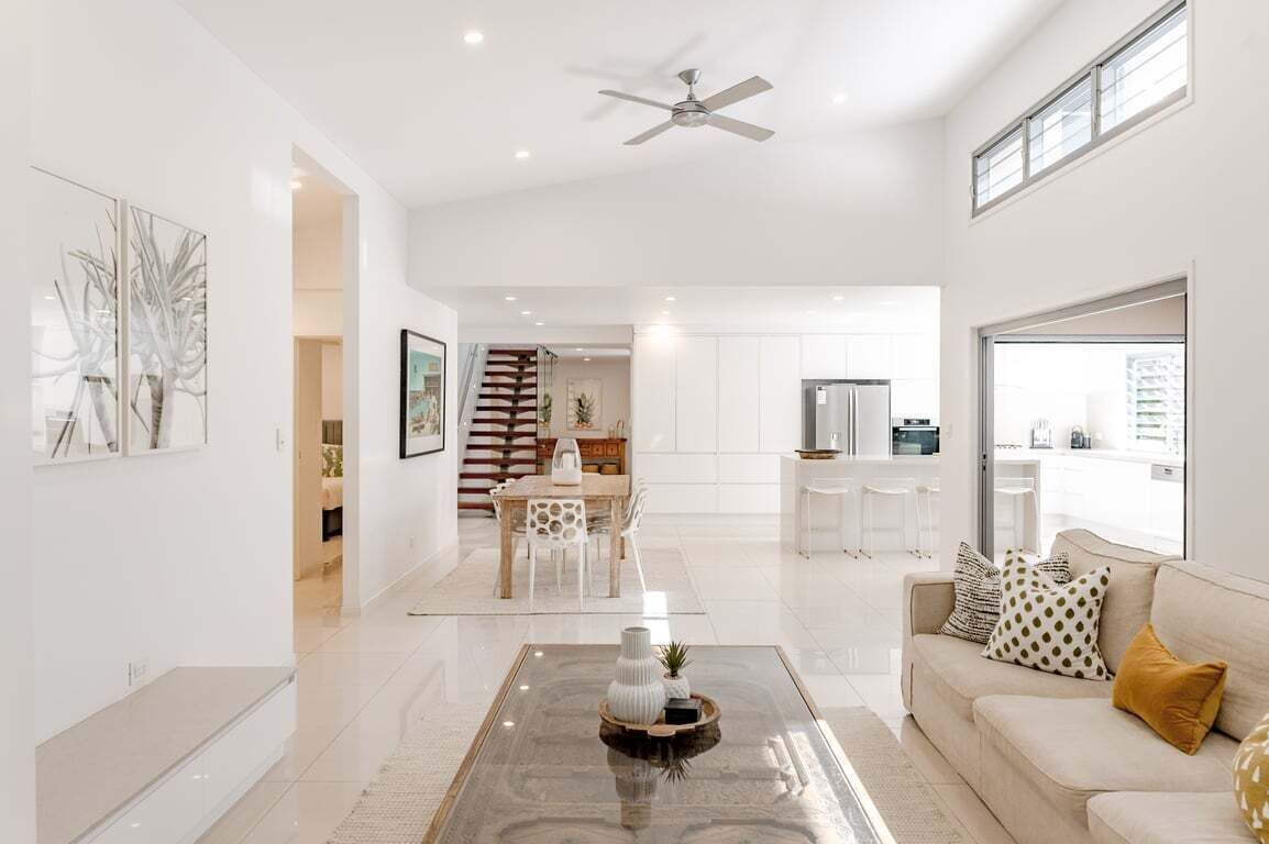A modern living room with a glass coffee table, a sofa with patterned cushions, a dining area with a wooden table and chairs, and a kitchen with white cabinets and a refrigerator.