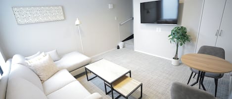Una sala de estar moderna con sofá, mesa de centro y sillas, un televisor de pantalla plana montado en la pared y una escalera que sube a un nivel superior.