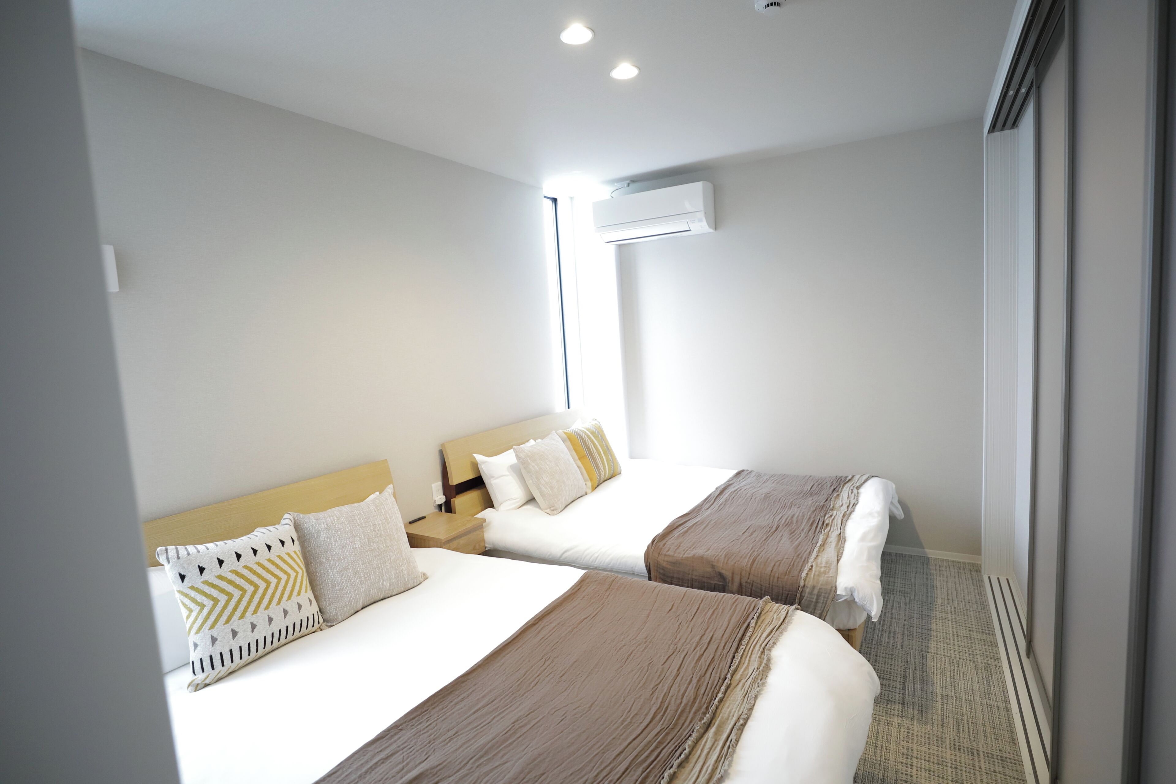 A hotel room with two beds, a window, an air conditioning unit, and a nightstand.