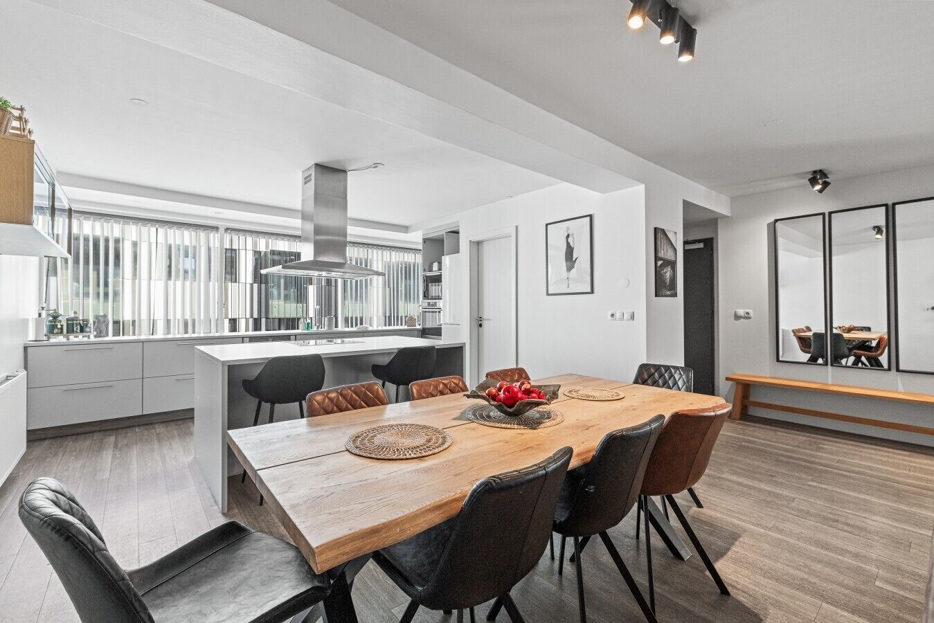 A modern kitchen with a central island, black chairs, and a wooden dining table with a bowl of fruit.
