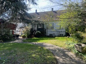 Un bâtiment doté d’un toit recouvert de tuiles, d’un chemin pavé et d’une zone herbeuse avec des arbres.